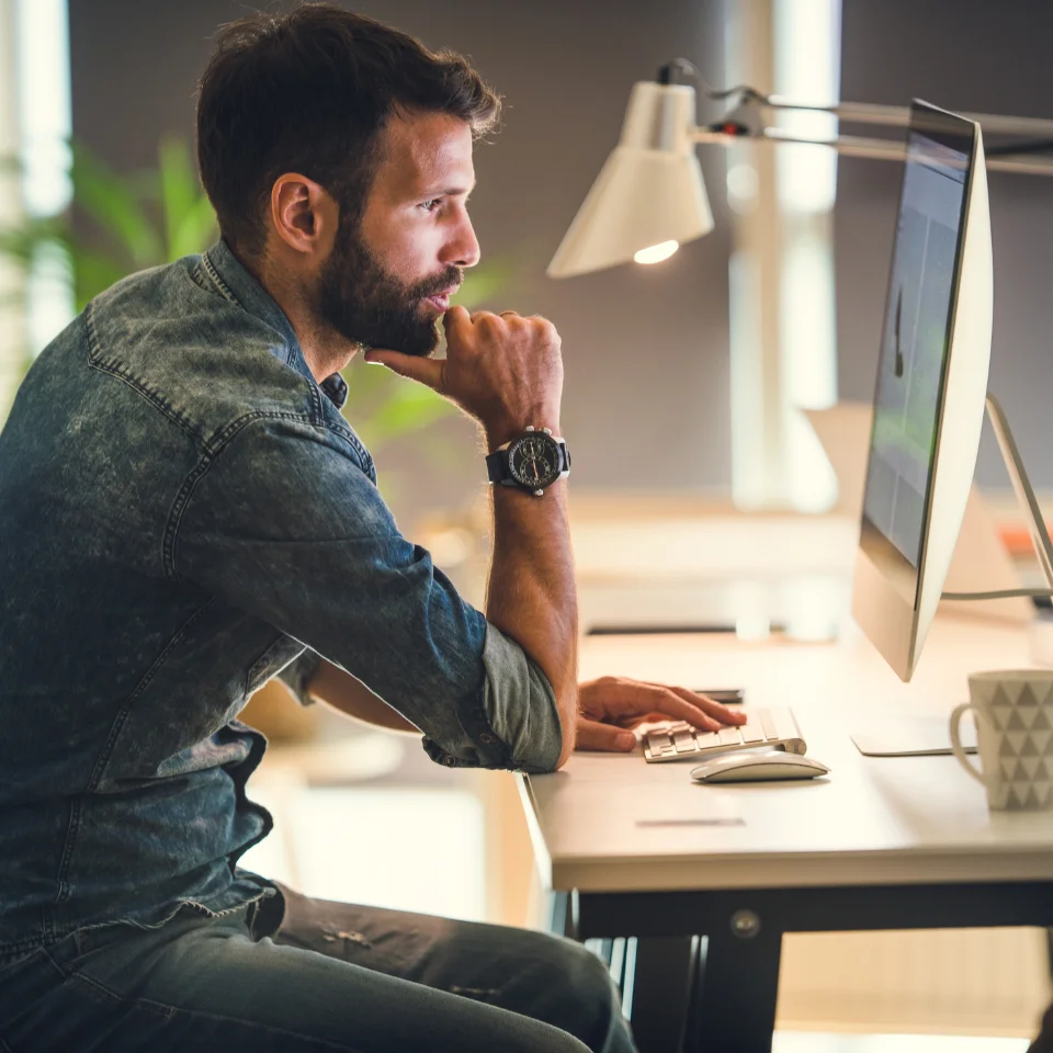 bearded-man-in-denim-shirt-working-at-computer-desk-with-hand-on-chin-in-thoughtful-pose
