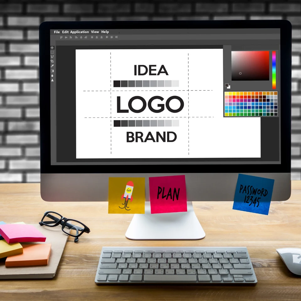 Computer screen displaying a logo design process with text 'IDEA', 'LOGO', 'BRAND' in a creative workspace with sticky notes