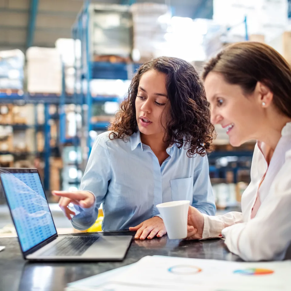 Two women discussing data on a laptop in a warehouse setting, collaborating on inventory or logistics while holding a coffee cup.