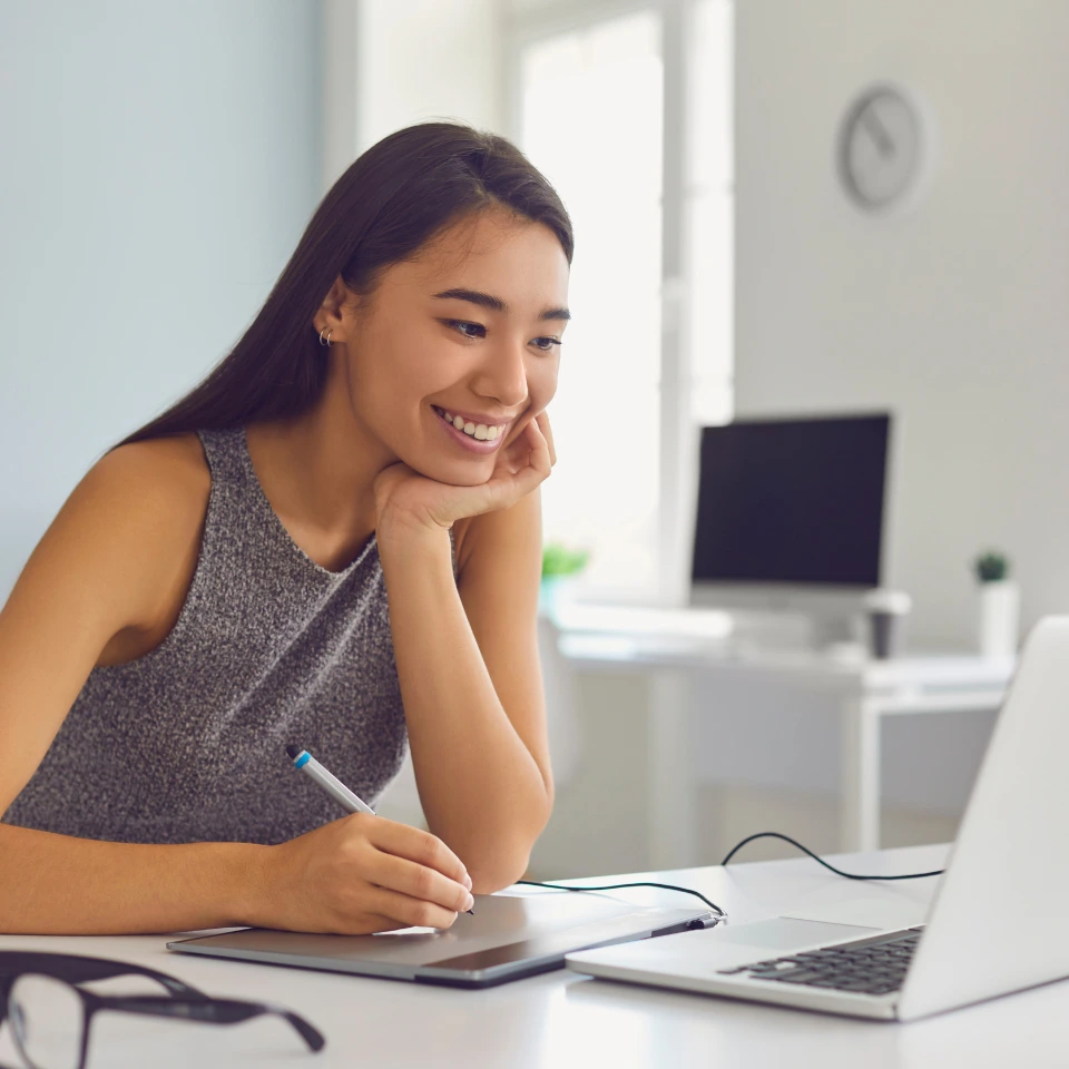 Smiling-young-woman-using-a-digital-tablet-while-looking-at-her-laptop-in-a-bright-modern-office-with-minimalistic-decor