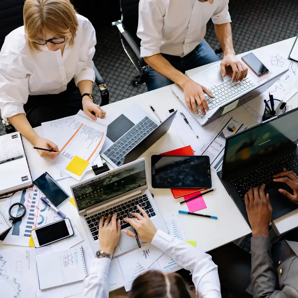 Group of business professionals working at a cluttered table filled with laptops, tablets, charts, and documents during a meeting.