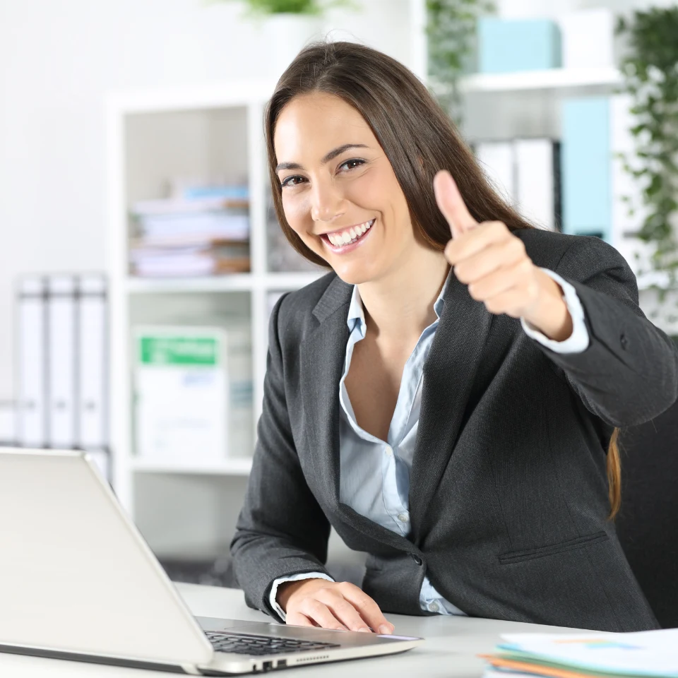 smiling-businesswoman-in-gray-suit-giving-thumbs-up-while-sitting-at-laptop-in-modern-office
