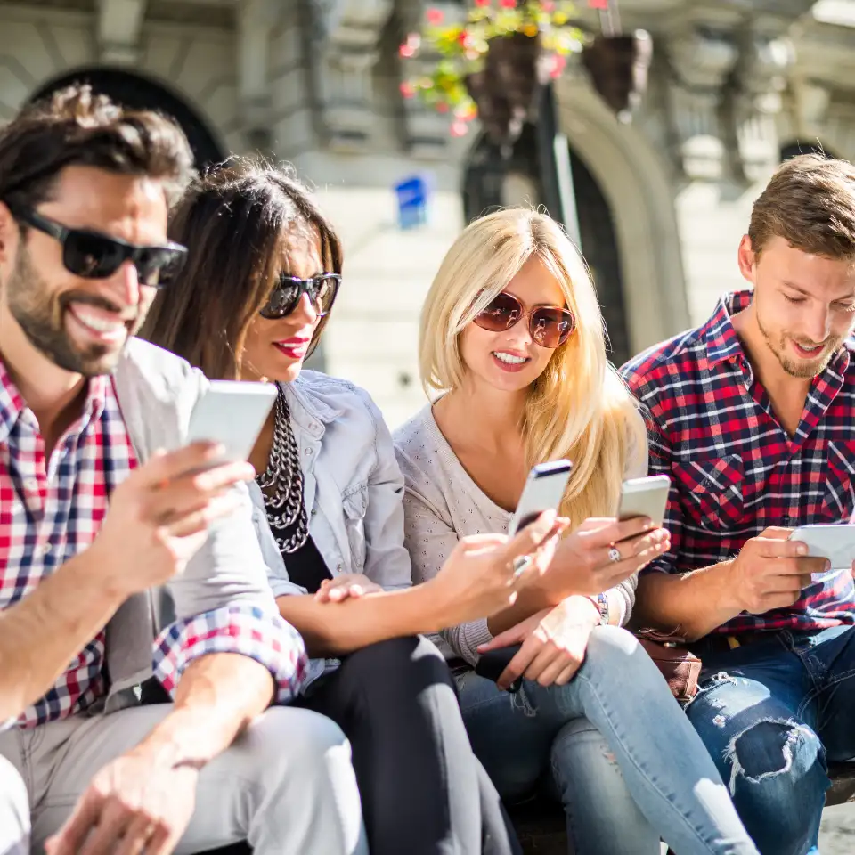 Group of young adults sitting outdoors, smiling and using smartphones, enjoying social media or online content on a sunny day