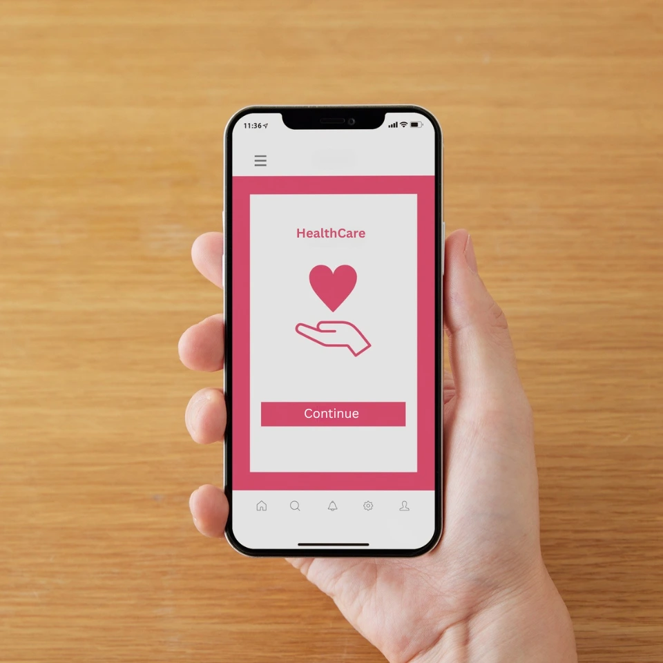 Healthcare professional in white coat holding smartphone with health app, laptop open showing medical dashboard on wooden desk.