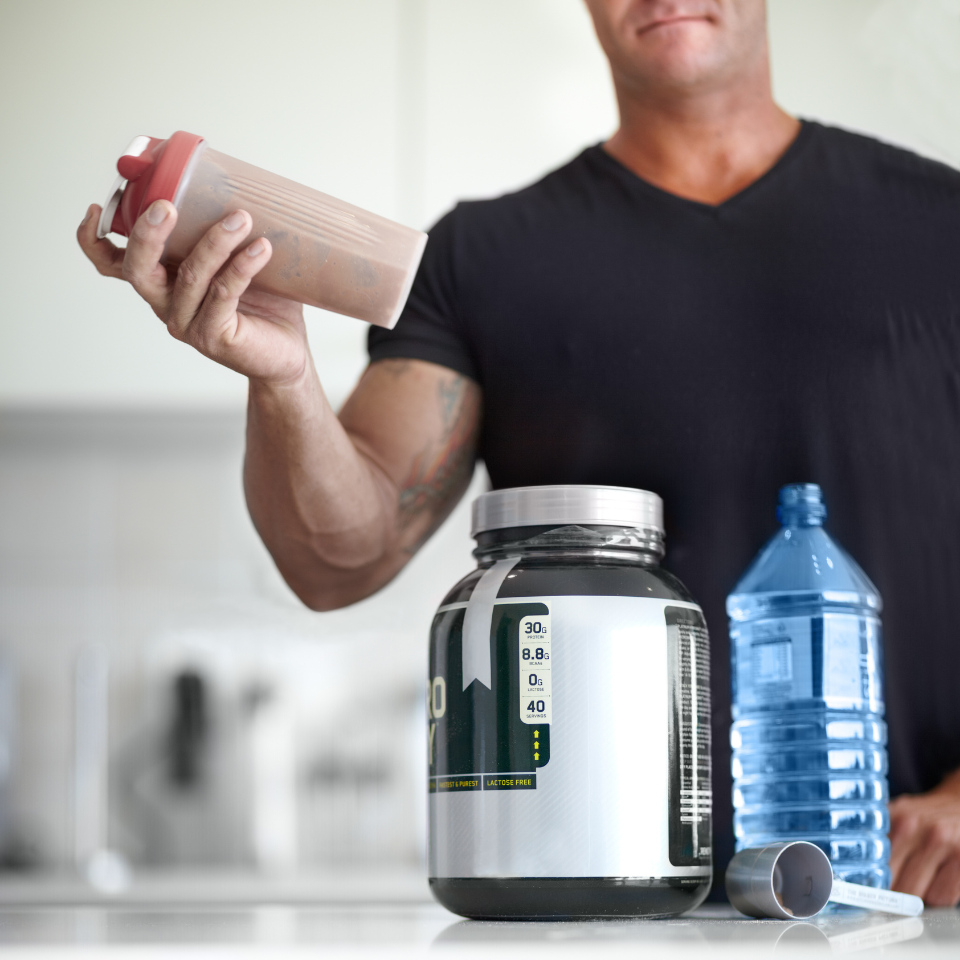 A muscular man in a black shirt holding a protein shake bottle, with a large protein supplement container and a blue water bottle on the kitchen counter in front of him.