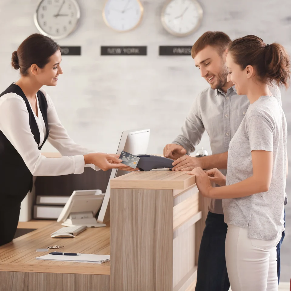 Smiling hotel receptionist handing key cards to a couple at the front desk during check-in, with clocks showing different time zones in the background.