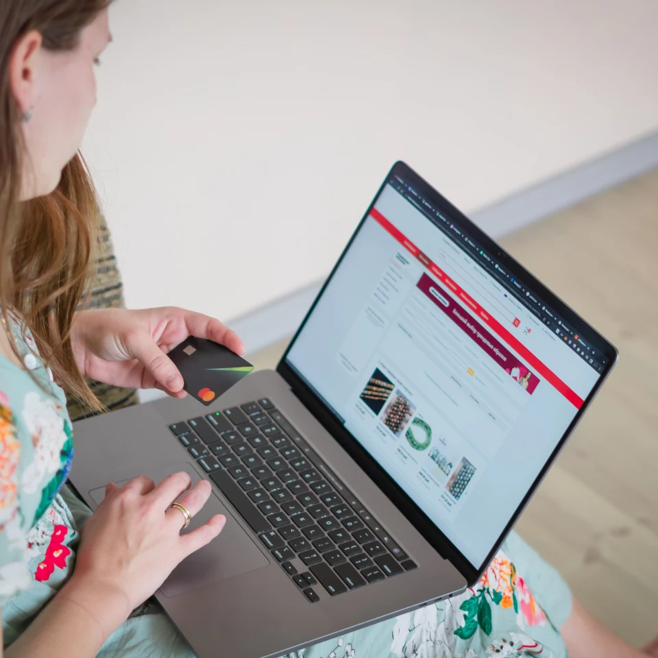 Woman holding a credit card while shopping online on a laptop, browsing an e-commerce website with product listings displayed on the screen.